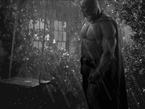 Batman looking sad as he pushes an empty child's swing in the rain