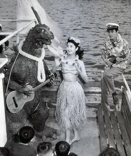 Godzilla playing ukulele with a Hawaiian lei around his neck. A Japanese hula girl dances beside him.