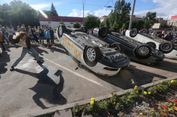 Three overturned cars surrounded by angry crowd at Russian embassy in Kiev, Ukraine. One protester throws a stone at an overturned, white, grafittied car.